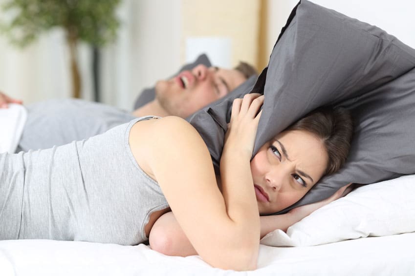 Eine Frau fühlt sich im Schlaf gestört durch ihren schnarchenden Partner und hält sich die Ohren mit einem Kissen zu